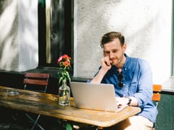 Junger Mann arbeitet mit Laptop an einem Holztisch im Freien vor einem Gebäude in der Sonne.